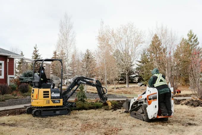 A landscaping crew clearing a yard with a mini excavator and a stand-on loader
